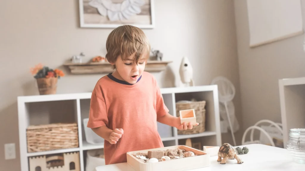generalização no autismo: menino observa uma caixa de madeira aberta com objetos dentro com cara surpresa e segura uma caixinha de madeira menor na mão esquerda