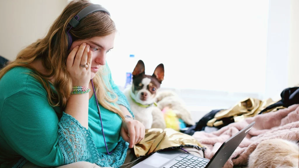 transtornos invasivos do desenvolvimento: menina está sentada com fones de ouvido e um notebook à frente. Um cachorro está observando ela na imagem.