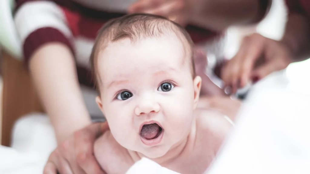 saúde: bebê com a boca aberta observa câmera, é possível ver uma mão o amparando