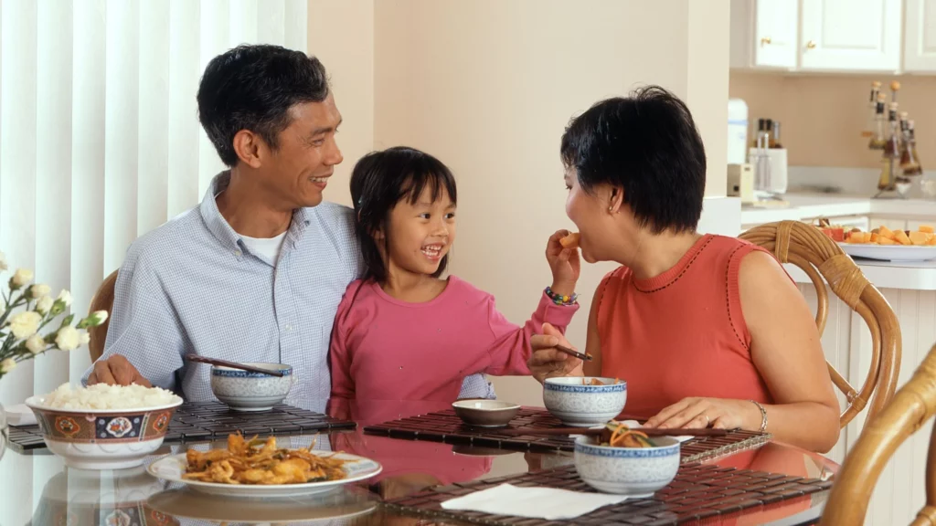 Lei Berenice Piana: mãe, pai e menina estão sentados à mesa e menina oferece comida na boca da mãe