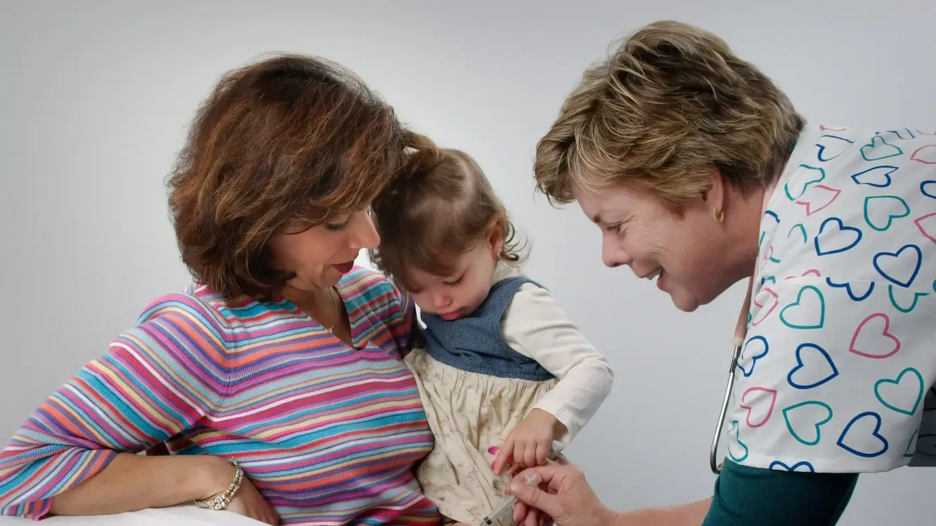 vacinas causam autismo? criança está sendo segurada no colo pela mãe e médica aplica injeção na perna dela