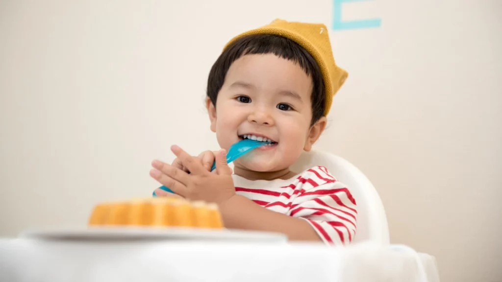 Criança está sentada com colher na boca, um bolo está a sua frente