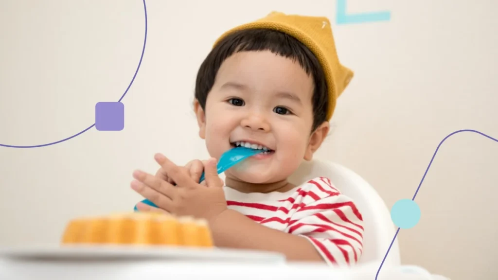 Criança está sentada com colher na boca, um bolo está a sua frente. Está desempenhando uma habilidade da vida diária no autismo