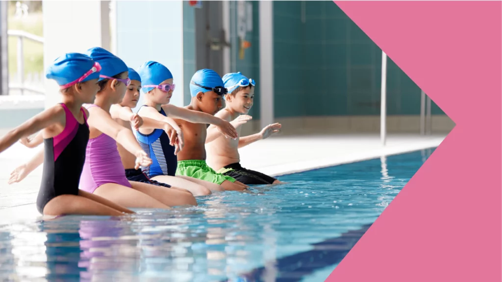5 crianças na beira de uma piscina prontas para uma aula de natação. todas estão com touca azul e roupa de piscina coloridas