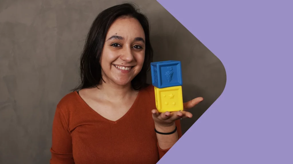 uma mulher sorrindo está segurando 2 blocos coloridos, um azul e o outro amarelo, na palma da sua mão.