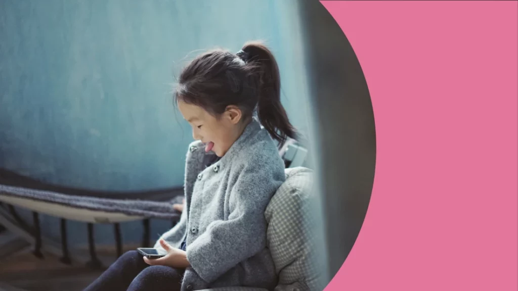 Menina asiática de cabelo preso e língua para fora, está sentada olhando a tela de um celular