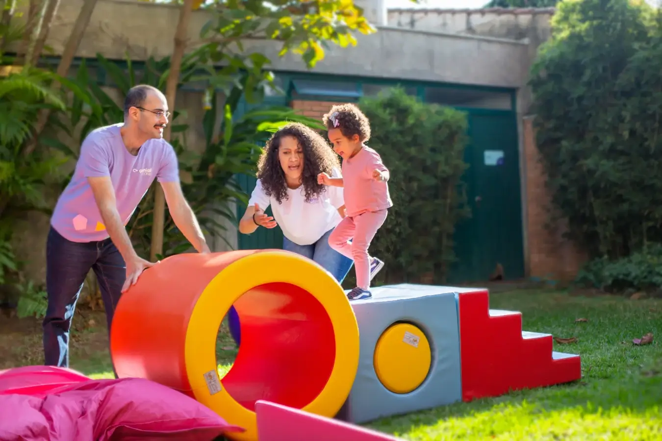 Terapeutas brincando com criança autismo. A brincadeira é feita sob brinquedos de obstáculos, em um jardim.