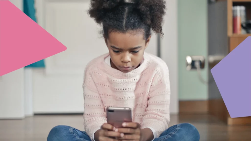 Menina negra com o cabelo preso está sentada no chão mexendo em um celular.
