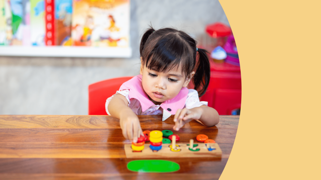 uma menina de cabelos pretos está sentada em uma mesa brincando com peças coloridas de encaixar em um bloco de madeira