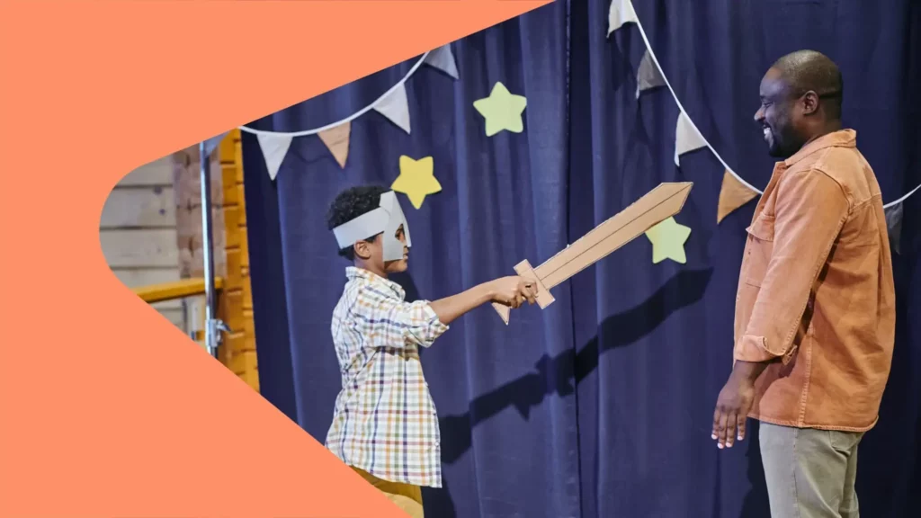 Atividades para autistas: Pai e filho estão em um palco de teatro, fazendo movimentos com uma espada falsa de madeira a fim de representar um personagem.