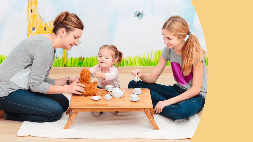 uma mulher de blusa cinza, uma criança de blusa rosa e uma adolescente de blusa cinza aparecem sentadas no chão em frente a uma mesa de madeira brincando de tomar chá