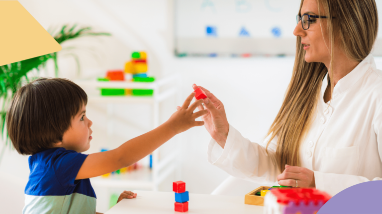 um menino de cabelo preto e blusa azul está pegando um bloco vermelho da mão de uma mulher loira de óculos. Ela testa mitos e verdade do autismo. Ambos estão sentados em uma mesa cheia de blocos coloridos.