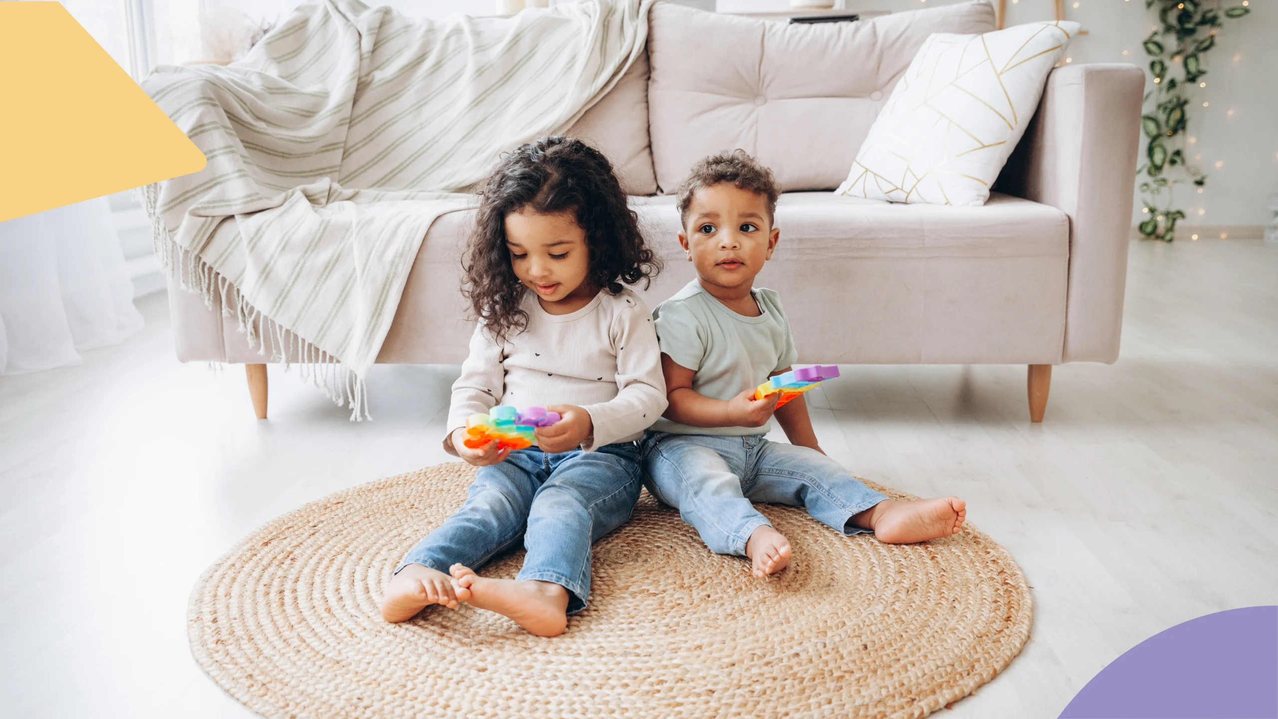 dois irmãos negros aparecem sentados no chão em cima de um tapete. ambos estão com brinquedos coloridos nas mãos