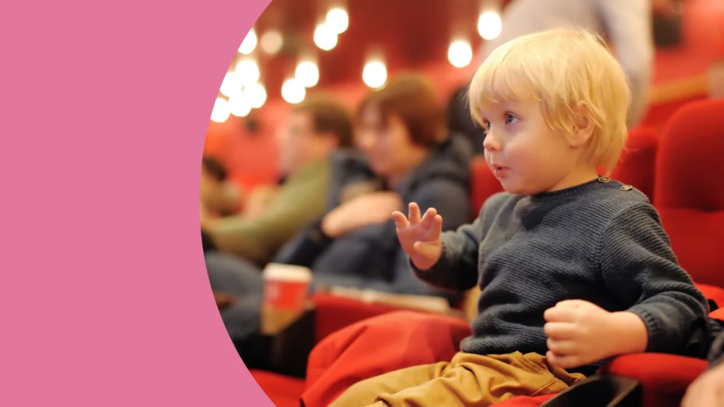um menino loiro aparece sentado em uma sala de cinema de poltronas vermelhas, e possível ver luzes acessas ao fundo