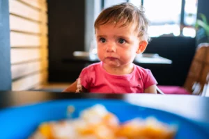 criança com comida na boca aparece séria em frente a um prato