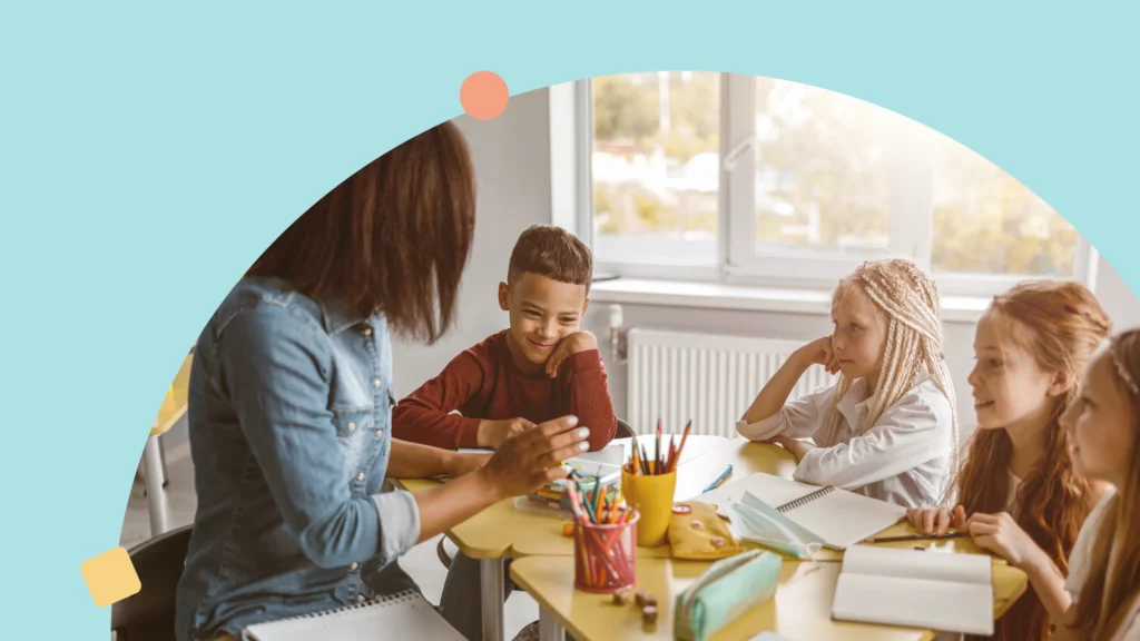 uma professora negra aparece sentada em uma mesa com 4 alunos, um menino negro e 3 meninas brancas. na mesa é possível ver vários materiais escolares