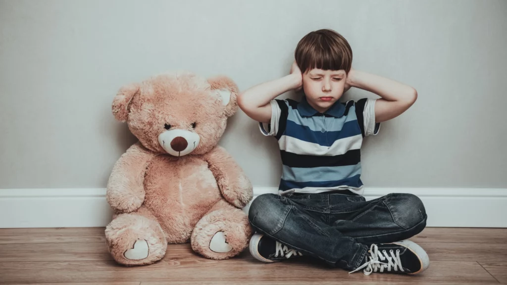 um menino de camiseta azul listrada em branco aparece sentado no chão tapando os ouvidos por ter sensibilidade auditiva, ao seu lado tem um urso de pelúcia marrom 