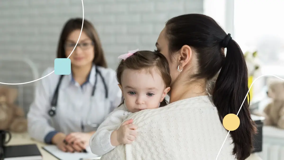 Mãe aparece abraçando um bebê que acabou de ser diagnosticado com autismo em um consultório médico, no fundo, é possível ver uma médica