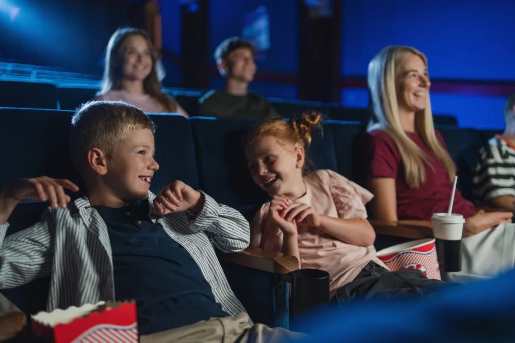 duas crianças aparecem rindo em uma fileira de cinema ao lado de uma mulher loira