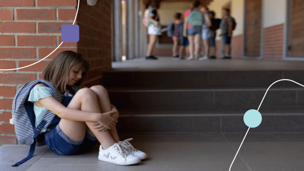 Menina sentada segurando as pernas. Ela está triste e ao fundo, tem várias crianças. O ambiente é escolar e representa recusa escolar