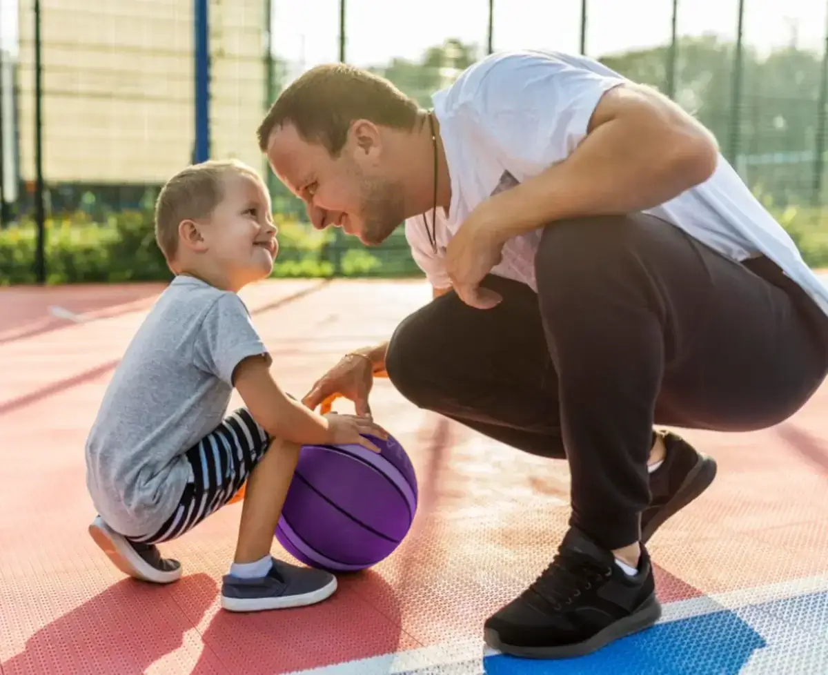 Pai e filho jogando Basquete. Ambos se oham agachados e seguram a bola com as mãos pressionando-a ao chão da quadra.