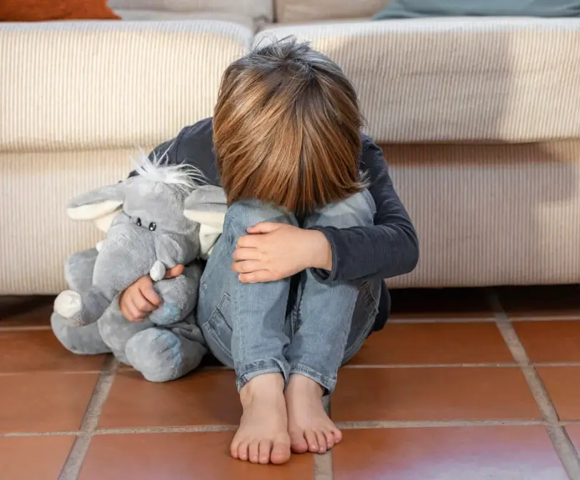 Menino sentado ao chão, abraçando seu urso de pelúcia. O menino apresenta Shutdown com o rosto entre os joelhos.