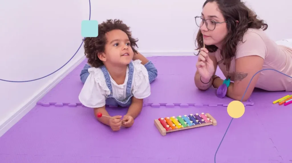 Terapeuta em carga horária de atendimento com criança autista. Ela senta deitada no chão de uma sala sensorial brincando com a menina.