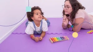Terapeuta em carga horária de atendimento com criança autista. Ela senta deitada no chão de uma sala sensorial brincando com a menina.