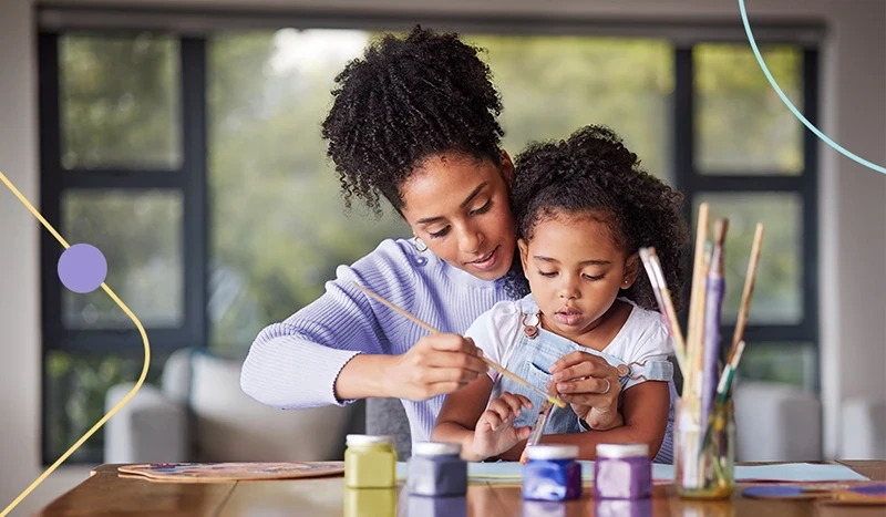 Mãe pintando desenhos com sua filha. Ambas brincam com tintas e pincéis.