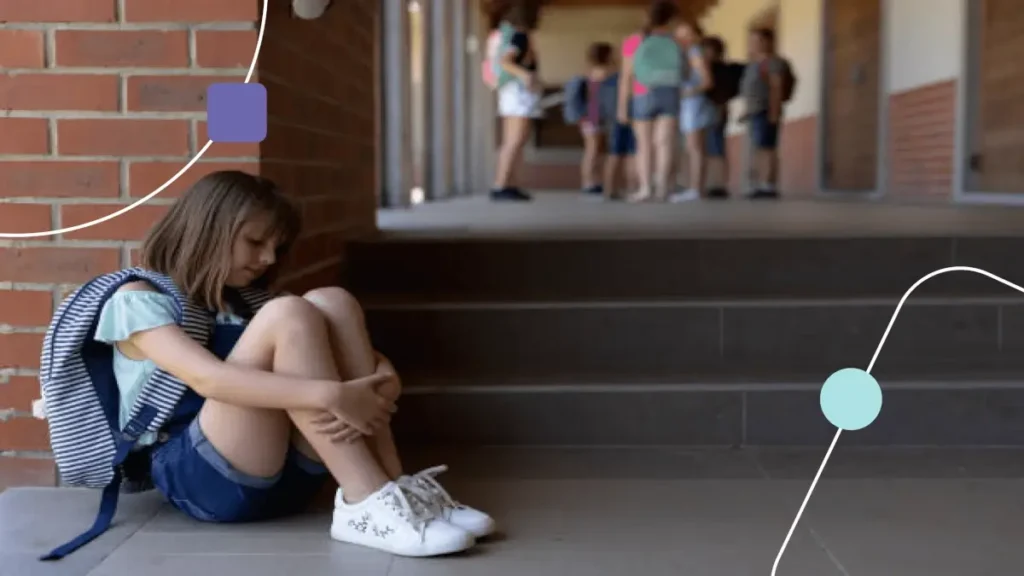 Menina sentada segurando as pernas. Ela está triste e ao fundo, tem várias crianças. O ambiente é escolar e representa recusa escolar