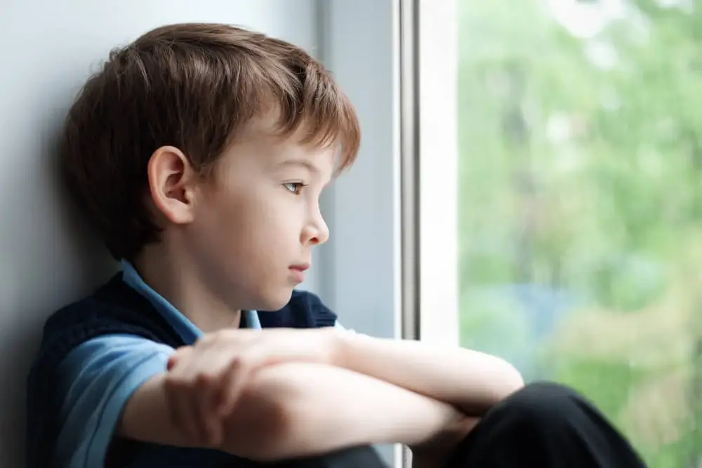 Menino triste olhando pela janela. Ele está sobrecarregado devido a diversas Atividades para autismo